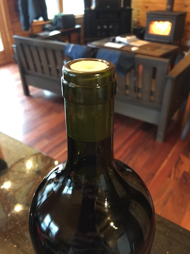 The top of a green wine bottle with a synthetic cork in it with a wood burning stove and a wooden couch in the background