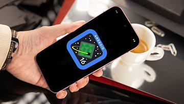 A hand holding a smartphone displaying the Android 16 logo over a table with a cup of coffee.