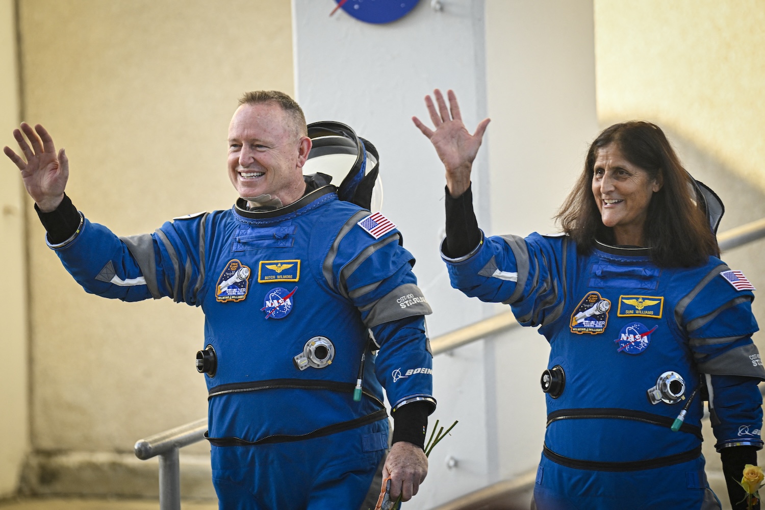 "NASA astronauts Butch Wilmore and Suni Williams, wearing Boeing spacesuits, wave as they prepare to depart."