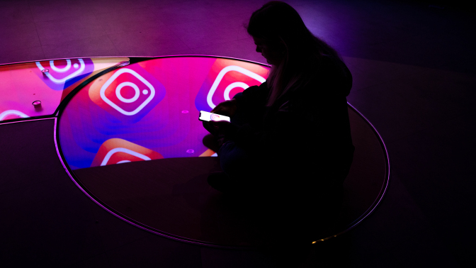 A person sitting in a dark room on their phone. A pattern showing the Instagram logo is reflected in mirrors below them. 