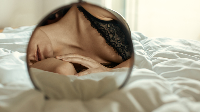 A woman lies on her side on her bed with a round mirror in front of her, her bra visible.