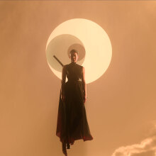 A woman with a sword floats in the air in front of three suns in an orange sky.