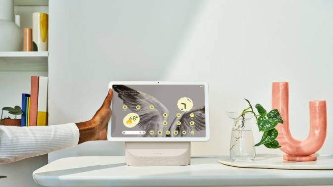 a google pixel tablet sits on a charging speaker dock on white tablet with a plant in a base beside it. 