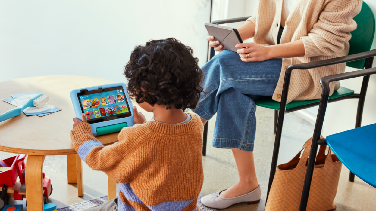 a toddler in an orange sweater plays with a blue fire 7 kids tablet while sitting at a wooden table. a woman in jeans and ballet flats uses a fire 7 tablet while sitting on a chair next to her