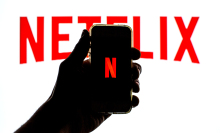 Silhouette of a hand holding a smartphone displaying the Netflix "N" in front of a white background with a large Netflix logo.