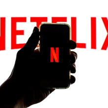 Silhouette of a hand holding a smartphone displaying the Netflix "N" in front of a white background with a large Netflix logo.