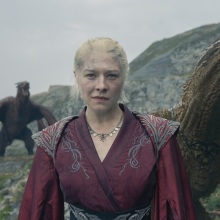 Rhaenyra stands on the island of Dragonstone, backed by two dragons.