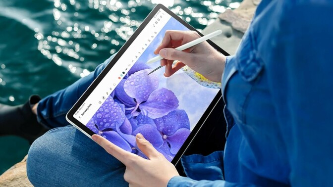 a person sits cross legged on some large rocks next to a body of water. They're using the included pen to draw on the  Samsung Galaxy Tab S9 FE+ tablet 