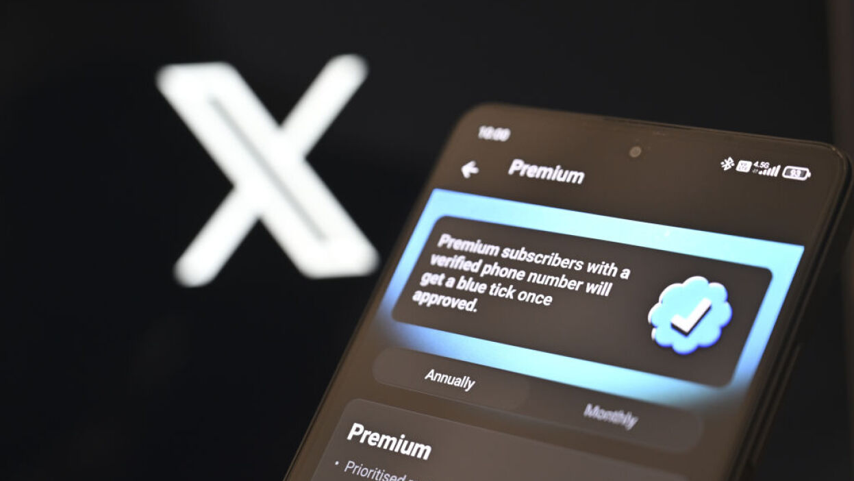 A page of 'X' (formerly known as Twitter) Premium account information is displayed on a mobile phone screen in front of a computer screen displaying 'X' logo.