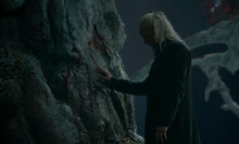 A man with long blonde hair stands in a dark courtyard, touching a tree.