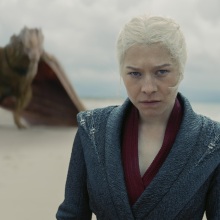 Rhaenyra Targaryen stands on a beach, with her yellow dragon Syrax behind her.