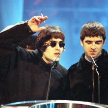 liam and noel gallagher on stage at an award ceremony. liam is gesturing with a cigarette in his hand, noel is looking downward.