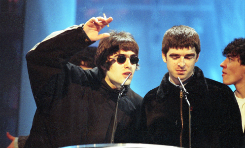 liam and noel gallagher on stage at an award ceremony. liam is gesturing with a cigarette in his hand, noel is looking downward.