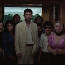 A group of men and women stand in a doorway, flanking a tall man in a white suit.