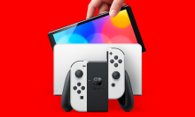 a person holding a nintendo switch oled model above its charging dock against a bright red background