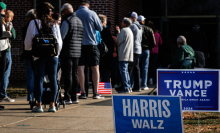 Harris and Trump yard signs are staked next to a line of voters entering a polling place.  