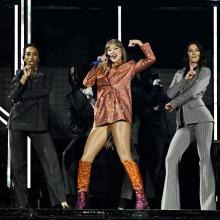 Taylor Swift performing in a blazer with one arm flexed as a demonstration of strength.