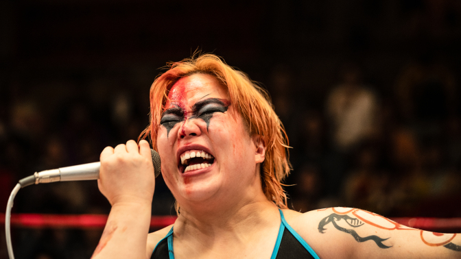 Professional wrestler with dramatic face paint and a microphone, passionately speaking during a match.