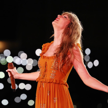 Taylor Swift in an orange dress looking up on stage. 