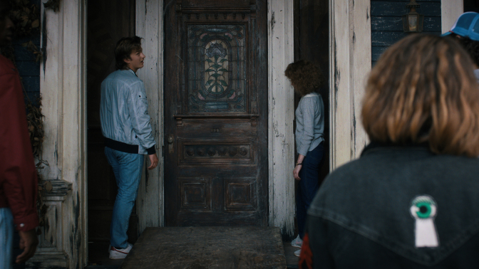 Teens stand outside a creepy old house door with a stained glass panel.