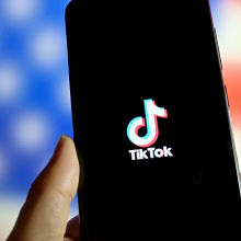 Person holds a phone showing the TikTok logo in front of a blurry American flag.