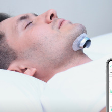 Person in white bed with device attached to chin and picture of phone showing data in front of them