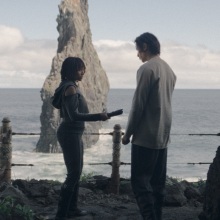 Osha holds an unlit lightsaber between her and the Stranger; the two stand at a cave entrance overlooking the ocean.