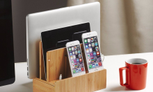 A Macbook, an iPad, and two iPhones sitting in a bamboo organizer and charging station next to a red coffee cup on a desk.