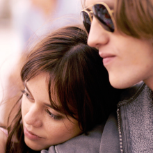 A young woman leans on the shoulder of a young man in sunglasses.