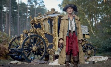 A young woman in a long coat and tricorn hat stands in front of a gilded carriage.