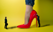 two tiny businessmen stand in front of a giant female foot in a red high heel in front of yellow background