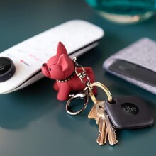 a side table includes a remote control, keys with a red dog keychain, and a wallet. All items have a tile bluetooth tracker attached.