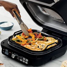 Person's hand using tongs to cook vegetables and chicken on Ninja griddle