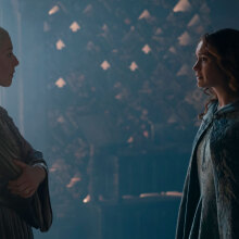 Two women in medieval clothes stand in a dark room, facing each other.