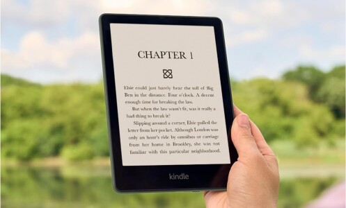 a person holds the amazon kindle in their right hand. The background has a forest scene with trees.