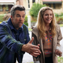 A man and a woman stand on the doorstep of a suburban road, looking excited.