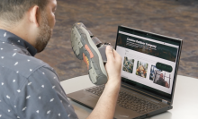 Man in blue shirt holding a hiking sandal in front of a computer