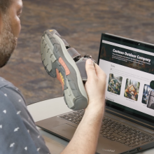 Man in blue shirt holding a hiking sandal in front of a computer