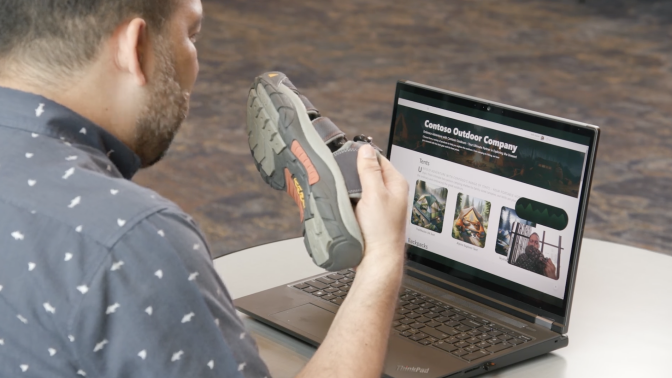 Man in blue shirt holding a hiking sandal in front of a computer