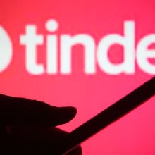 A red backdrop including the Tinder logo with the silhouette of a person's hand holding a phone. 