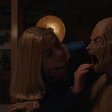 A woman reaches her hand into the mouth of a wooden man. 