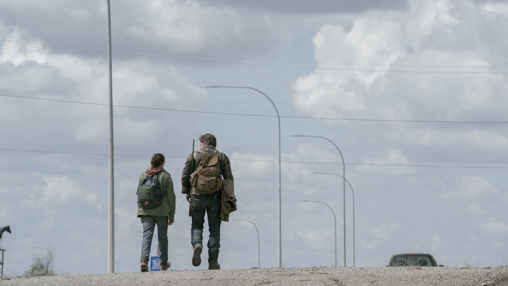 A man and a teen wearing backpacks and carrying guns walk down an abandoned road.