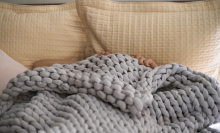 A woman lies under a knitted blanket in bed with just her hands visible. 