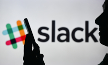 A woman holding a cell phone in front of the Slack logo displayed on a computer screen.