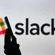 A woman holding a cell phone in front of the Slack logo displayed on a computer screen.