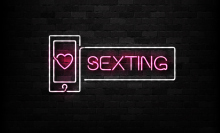 A neon sign with pink text which reads "sexting" on a black brick wall.