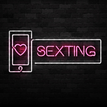 A neon sign with pink text which reads "sexting" on a black brick wall.