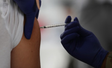 A gloved hand injects a needle into a person's upper arm.