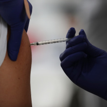A gloved hand injects a needle into a person's upper arm.