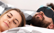 Person wearing black sleeping mask sleeping next to smiling person with eyes closed
